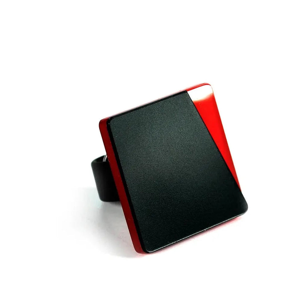 A square-shaped Saylor Ring with a black and red geometric design on its top, crafted from high-quality acrylic glass by Alex + Svet. The ring's surface is predominantly black with a red accent on one corner, creating a sleek and modern look. The band is also black and visible from the side. The background is white.