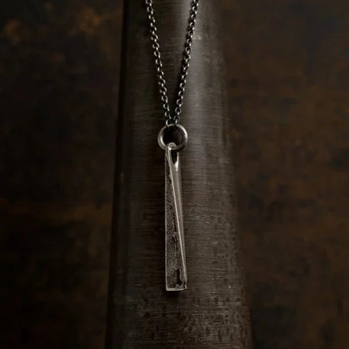 A Large Tetrahedron Necklace crafted from Patinated 925 silver with a rough texture hangs from a dark chain necklace. This piece of handmade jewelry by Septentrion is positioned against a dark, rustic background.