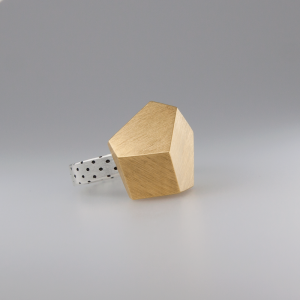 A gold, geometric-shaped Fruit Bijoux Ring Top Goldrush with an angular design and a textured surface. The band, crafted from 925 sterling silver with small black dots, showcases the beauty of handmade jewellery by Fruit Bijoux. The ring is displayed against a smooth, light gray background.