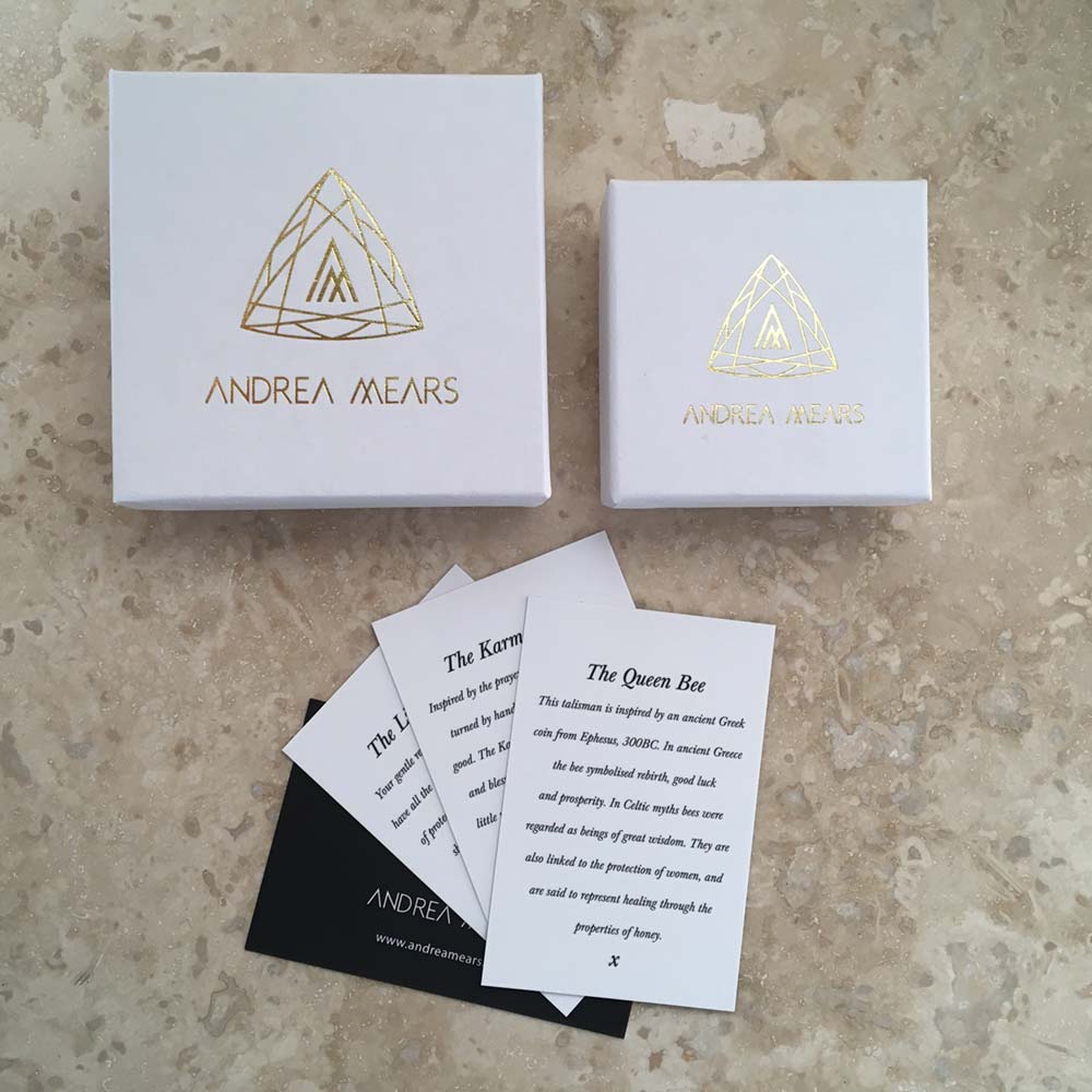 Two jewelry boxes branded "Andrea Mears Jewellery" are on a marble surface. The larger box is open, its lid leaning against it, revealing information cards inside. These cards describe various sterling silver pieces, including the Karma Wheel Necklace Silver inspired by Buddhist Tibet and "The Queen Bee.