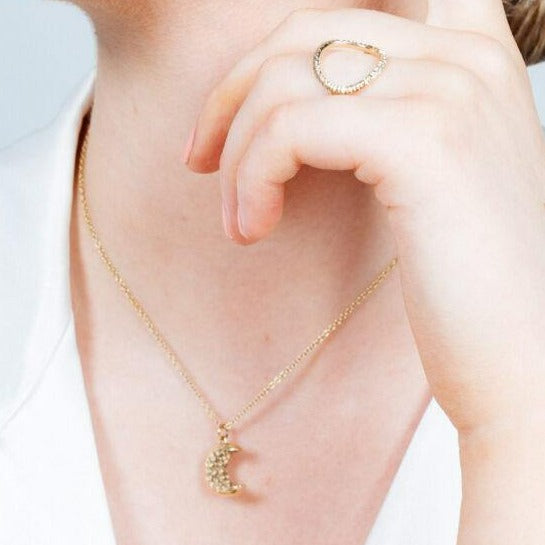 Close-up of a person wearing a gold necklace with a crescent moon pendant and a textured circle ring on their finger. The Into The Light Ring 9ct Yellow Gold by Aud gleams against their white top, as only their neck and lower face are visible while their hand gently touches their chin.