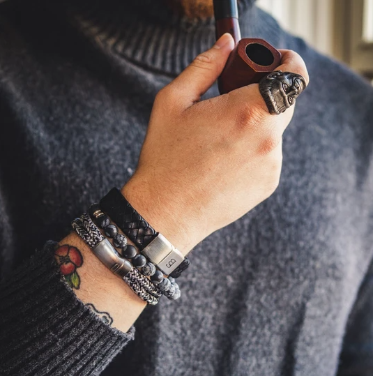 A person wearing multiple bracelets with stainless steel clasps and a ring, holding a wooden pipe. Their arm has a tattoo of a red flower with green leaves, and they are dressed in a grey knit sweater featuring the Preston - Matt Black bracelet by Steel and Barnett.