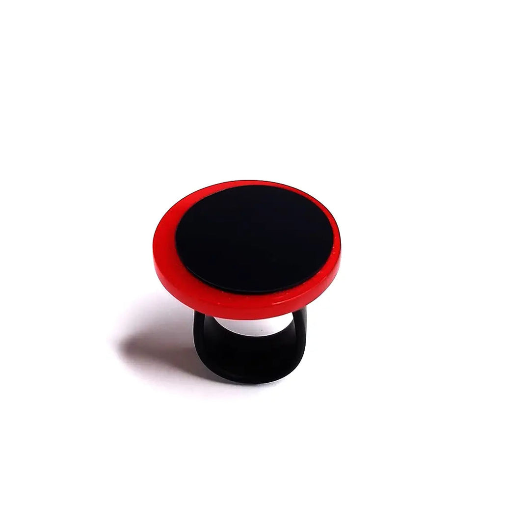 An Alex + Svet Amy Ring stands against a plain white background. The top surface is black, while the outer edge is red. Resembling a statement ring, the grip is designed to attach to the back of a smartphone for improved handling and support.