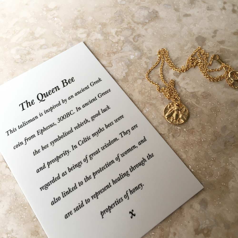 A Queen Bee Necklace Silver from Andrea Mears Jewellery is displayed next to a card on a light textured surface. The card is titled "The Queen Bee." Text on the card explains the historical significance of bees in Greek and Celtic cultures and their symbolism of wisdom, protection, and healing.