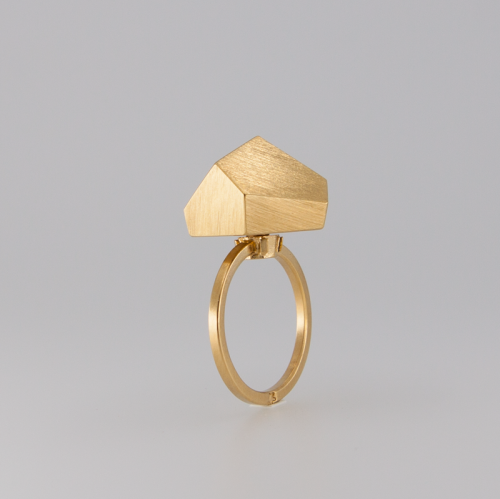 The Fruit Bijoux Ring Top Goldrush by Fruit Bijoux features a unique house-shaped structure on top, with a smooth, polished band and a matte finish on the house. This piece of handmade jewelry is centered in the image against a light gray background, standing upright.