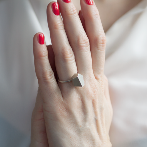 Close-up of hands with red-painted nails clasped together, showcasing a Fruit Bijoux Ring Top Goldrush by Fruit Bijoux. The background features a person in a white, blurred garment.