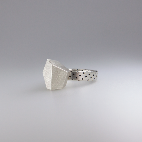 A **Fruit Bijoux Ring Top Goldrush by Fruit Bijoux** with a polygon-shaped, textured top and a band featuring a pattern of small, evenly spaced black dots. This piece of handmade jewellery is displayed on a smooth, light gray surface with a gradient background.