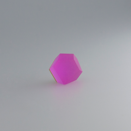 A small, transparent, faceted geometric object with a pink hue sits on a plain, light-gray surface. The object, likely a Fruit Bijoux Ring Top VU Crystals Magenta Pink by Fruit Bijoux destined for a sterling silver ring top, is illuminated to emphasize its clear, vivid color and precise shape.