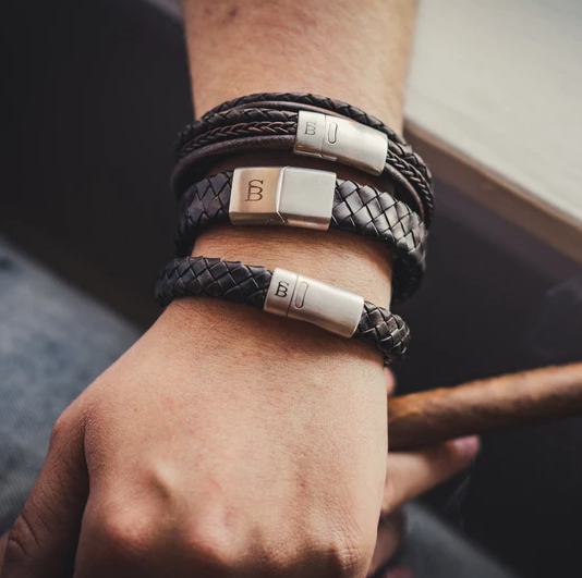 A person's wrist is adorned with three Steel and Barnett Preston - Vintage Brown braided leather bracelets, each featuring a metallic clasp. The person holds a cigar in their hand, exuding a masculine accessory vibe. The background appears to be a blurred, indoor setting.