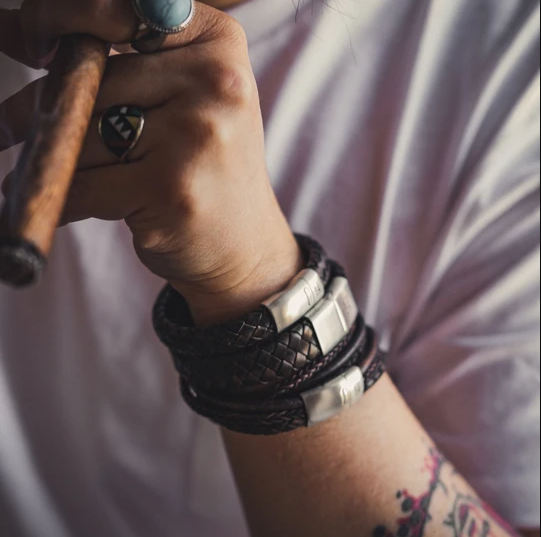 A close-up of a person holding a cigar in their left hand, adorned with masculine accessories like multiple leather bracelets and silver rings, including one with a blue stone. Genuine leather details and visible tattoos complete the look against the blurred background, showcasing their white shirt. Among the accessories is the Preston - Vintage Brown bracelet by Steel and Barnett.