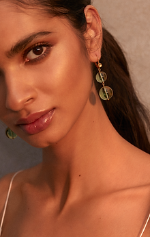 A close-up of a woman with long dark hair tied back, wearing elegant Pernille Earrings by Shyla. She has warm-toned makeup with a gold highlight on her cheekbones and neutral lip gloss. She is wearing a thin-strapped top and looking directly at the camera.