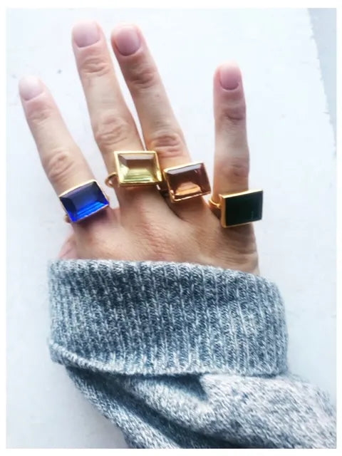 A hand adorned with four large, rectangular Lenny Rings by Shyla of various colors. The rings, crafted from lustrous 22-carat gold, feature stunning blue, yellow, pink, and black stones. The person is elegantly dressed in a gray sweater.