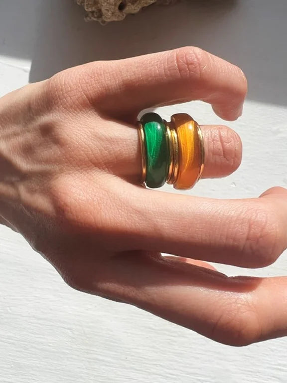 Close-up of a hand with two chunky rings on the middle finger. One gold band boasts a green stone, while the other features an amber stone. Both stones are shaped like smooth, rounded cabochons. This Orion Ring by Shyla is beautifully illuminated by natural sunlight.