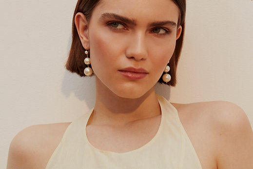 A woman with short brown hair wears a sleeveless cream top and elegant Shyla Pernille Earrings. She looks slightly to the side with a thoughtful expression. The background is a plain white wall.