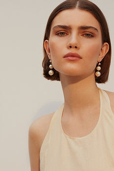 A woman with short brown hair and a serious expression wears a sleeveless, light-colored top. She accessorizes with elegant Pernille Earrings by Shyla. The background is plain and softly lit, highlighting her face and outfit.