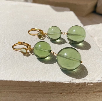 Introducing the Shyla Pernille Earrings: a pair of elegant dangle earrings featuring two green translucent glass beads of varying sizes on each earring. The beads are connected to 22 carat gold hooks with a smaller green bead near the clasp. These stunning earrings rest on a light, textured surface.