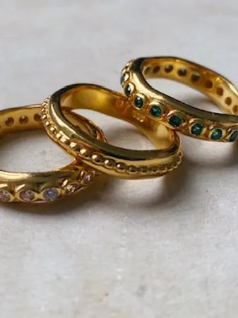 Three Shyla Astri Stacking Rings in 22 carat gold are arranged on a flat surface. Each ring features different embellishments: one with small diamonds, another with tiny green gemstones, and the last with a textured pattern. These hypoallergenic treasures combine beauty and comfort perfectly.