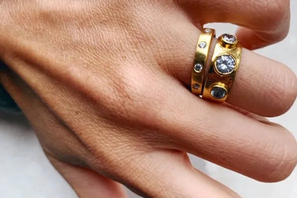 A hand adorned with two 22 carat gold rings on the ring finger. The top ring, possibly a Shyla Mela Ring, features a large central gemstone and smaller gemstones around it, while the bottom ring has small evenly spaced gemstones embedded around it. The hand is shown against a light-colored background.