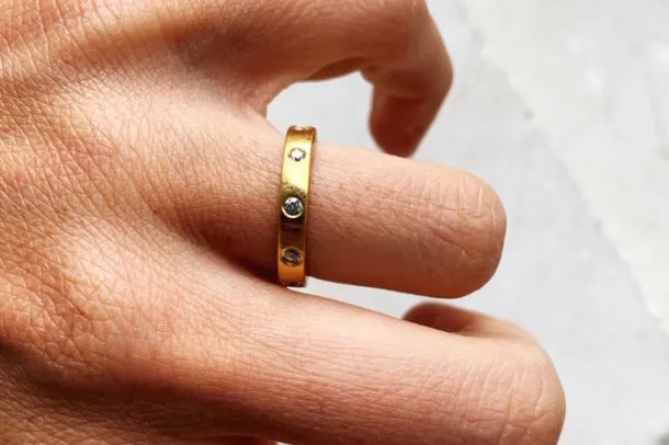 A close-up of a hand wearing a Shyla Mela Ring in 22 carat gold with small, embedded gemstones. The skin tone is light, and the background is blurred, highlighting the ring's details. The ring appears to be simple yet elegant with its minimalistic design.