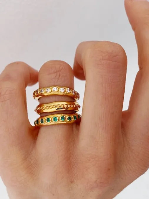 A hand with four fingers adorned with Shyla Astri Stacking Rings made of 22 carat gold. The rings on the middle finger are decorated with different gemstones: white and green. The background is a plain, light-colored surface, creating an elegant and hypoallergenic display.
