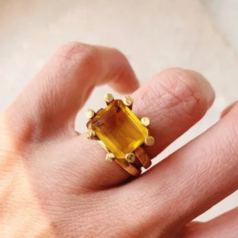 A hand wearing a Square Claw Gold Plated Ring from Shyla. The ring features a large, square yellow gemstone securely held by multiple gold prongs, and the 22 carat gold ring boasts a unique and elegant design. The background is softly blurred.