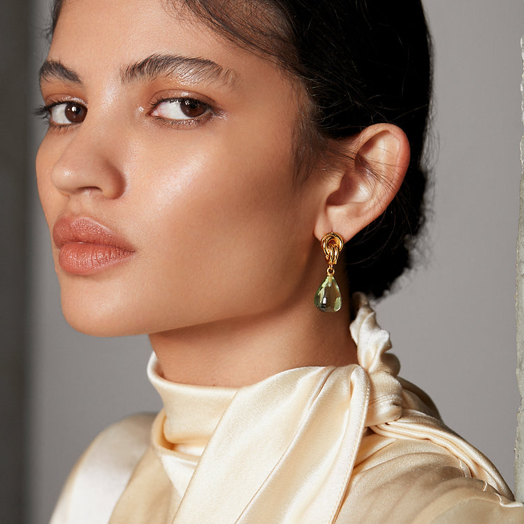 A person with smooth, glowing skin and neatly tied-back dark hair looks confidently into the distance. They are adorned with Shyla's Chunky Knot Earrings in high shine gold featuring a large green teardrop gem and are wearing a silky, cream-colored top with a stylish scarf-like collar.
