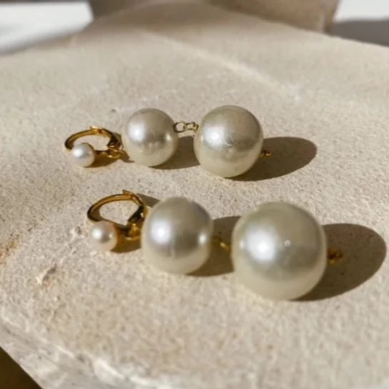 A pair of Pernille Earrings by Shyla in 22 carat gold, adorned with spherical white pearls, are shown on a textured, light-colored surface. Each elegant earring features two pearls of different sizes, the larger pearl hanging below the smaller one. The earrings are bathed in natural sunlight.