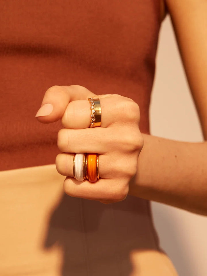 A person wearing a sleeveless brown top and beige bottoms shows their hand adorned with three chunky rings: a thick gold band, a delicate chain design, and one featuring an orange stone. Their hand is closed in a fist, with the hypoallergenic Shyla Orion Ring prominently displayed.