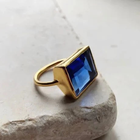 A 22 carat gold ring featuring a large, rectangular blue gemstone is displayed on a light-colored stone surface. The gemstone is cut with sharp, clean facets, highlighting its deep blue hue, making the Shyla Lenny Ring a stunning cocktail ring and true statement piece.