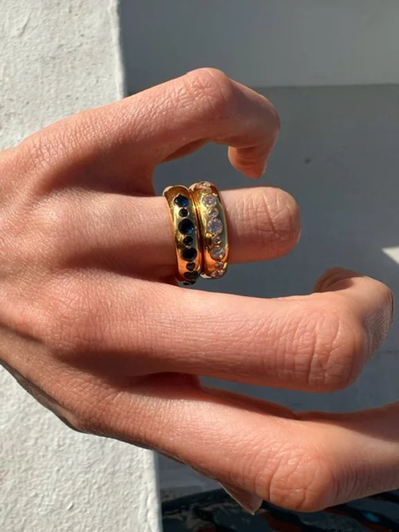 A close-up of a hand with two fingers adorned with 22 carat gold rings. One ring, potentially a Tasha Ring by Shyla, features a row of black gemstones, while the other displays colorful jewels. The hand is positioned against a light-colored wall with visible shadows.