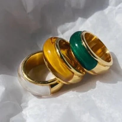 A close-up photo of three chunky Orion Rings by Shyla with gold-colored bases and different enamel inlays. The hypoallergenic jewelry features inlays in white, yellow, and green, and is arranged side by side on a crumpled, light-colored background.