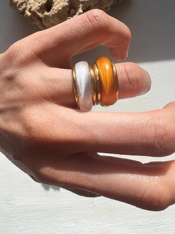 A hand wearing three chunky rings on the index finger: one in silver, one in gold, and one in copper. The hand rests on a white surface with a textured beige object in the background. The warm lighting highlights the metallic finish of these Orion Rings by Shyla.