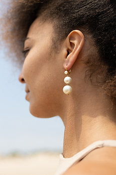 A close-up side profile of a person with curly hair wearing elegant Shyla Pernille Earrings. The 22 carat gold double pearl drop earrings consist of two stacked pearls hanging from a small, delicate hook. The background is blurred, with subtle shades of blue and beige.