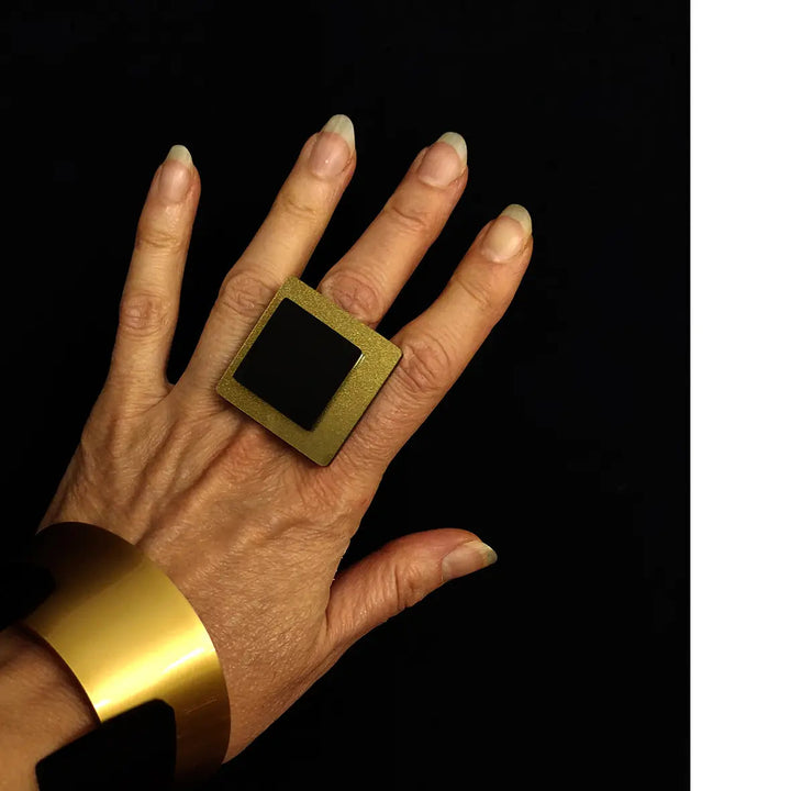 A hand with long, natural nails is adorned with a large, square Eriko Ring by Alex + Svet on the middle finger and a wide, gold bracelet on the wrist, set against a black background. This contemporary touch is part of the INFINITE LINE collection.