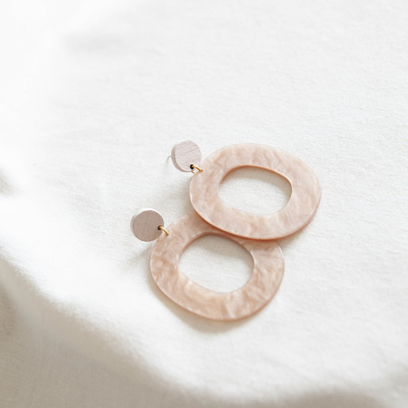 A pair of hand made, beige oval-shaped Studio Nok Nok Acrylic 09 Earrings with a marble-like texture, each featuring a small round stud at the top. Gold plated and crafted from upcycled ash wood, they rest on a white fabric surface with soft lighting highlighting their texture and color.