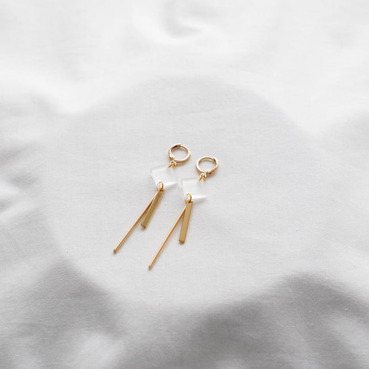 A pair of Glow 02 Gold Earrings by Studio Nok Nok laid out on a soft, white fabric. Each earring features a delicate geometric design with a combination of rectangular and elongated shapes, adding a touch of modern elegance to these handmade jewelry pieces.