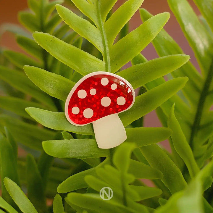 A vibrant red Mushroom Pin Brooch by Naoi with white polka dots, designed to resemble a vinyl mushroom, rests on lush green fern leaves. The pin's stem is white and the backdrop provides a natural setting for this striking accessory, complete with brass butterfly pin clasps.