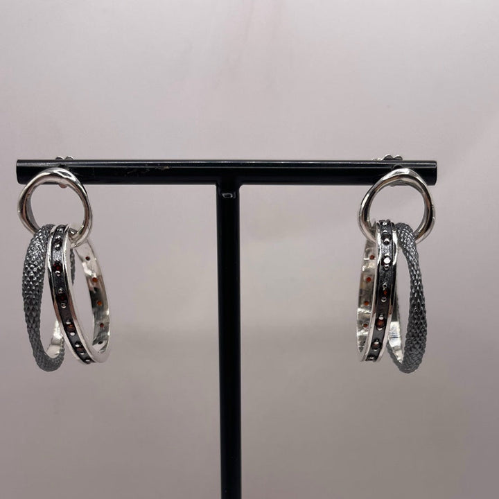 The K.And Double Circle Oxidised Earrings with Red CZ feature two interlocking textured and smooth bands, shown on a black stand against a plain light background.