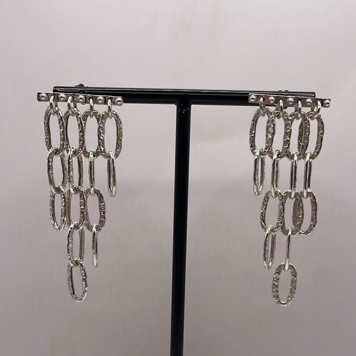 K.And Oval Chain Link Dangle Earrings in Silver or Oxidised feature multiple textured oval links hanging in three vertical rows, showcased on a black display stand against a plain light background.
