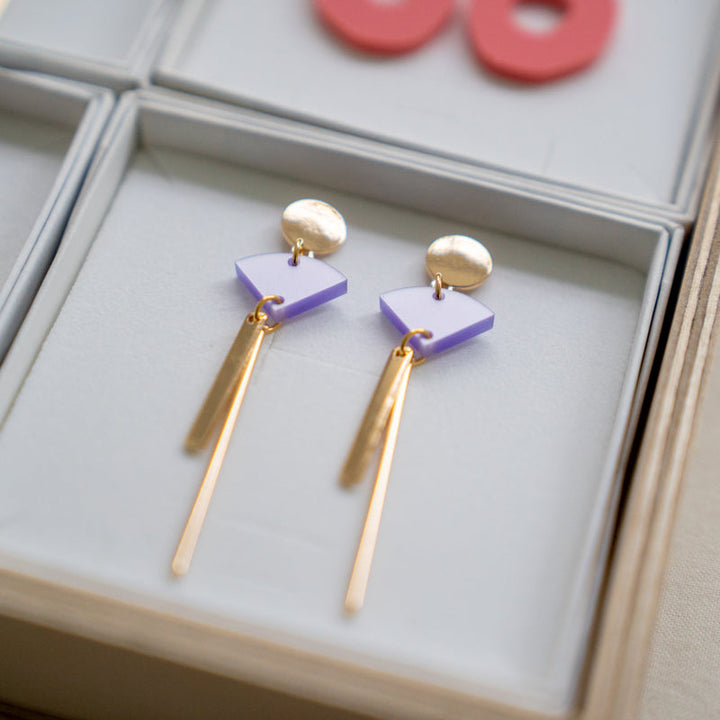 A pair of elegant Sun 08 Earrings displayed in a jewelry box. Each earring, crafted by Studio Nok Nok, features a gold circular stud with a purple, diamond-shaped gem beneath it and two slender, dangling gold rods extending downwards. The box has a padded interior for secure placement.