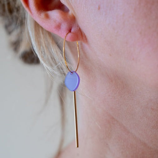 Close-up of a woman's ear wearing the Smile 06 Earrings by Studio Nok Nok, featuring a gold hoop with a purple circular pendant made from recycled acrylic, and a long, thin gold bar dangling from it. The woman's light blonde hair is partially visible against the slightly blurred background.