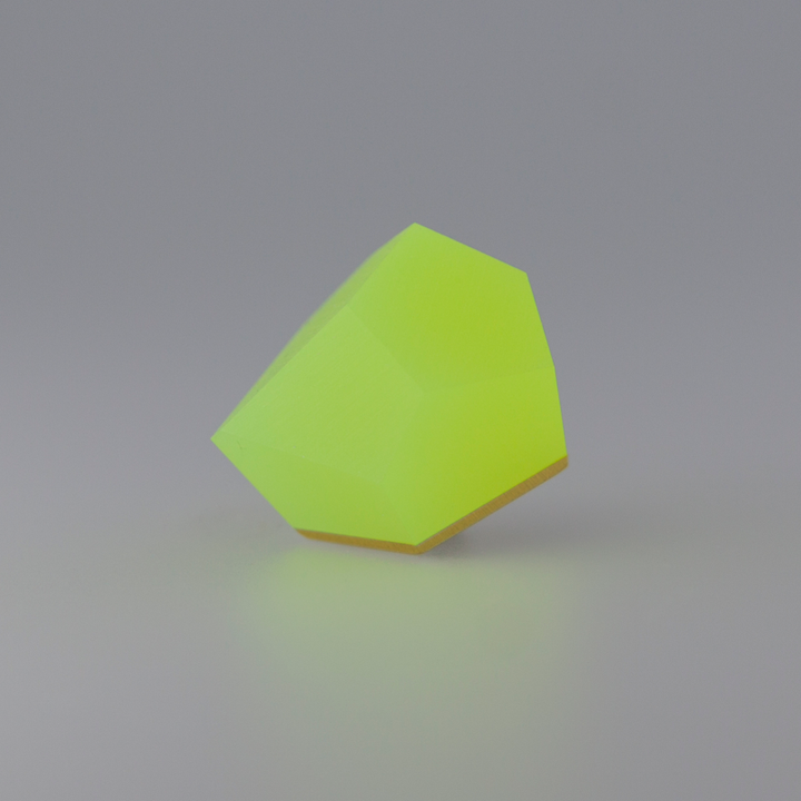 A bright yellow geometric object with multiple flat surfaces resembling a gemstone is placed on a smooth gray background. The Fruit Bijoux Fruit Bijoux Ring Top VU Crystals Neon Yellow, crafted with 24ct gold-plated elegance, has a slightly pointed end and a flat base, creating an abstract, modern design.