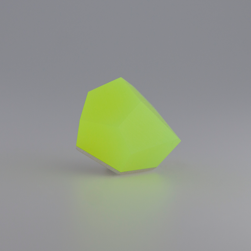 A brightly glowing, faceted gemstone with a yellow-green hue is displayed against a soft grey background. The gemstone's surfaces are smooth and angular, reflecting the light in various directions, and it is set in a sterling silver Fruit Bijoux Fruit Bijoux Ring Top VU Crystals Neon Yellow.