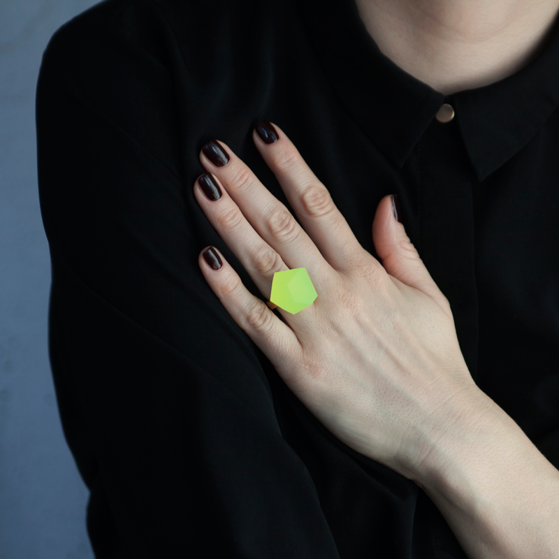 A person wears a black shirt with a gold button at the collar and rests their hand on their chest, displaying a bright yellow geometric Fruit Bijoux Ring Top VU Crystals Neon Yellow by Fruit Bijoux. Their nails are painted a dark shade, complementing the outfit.