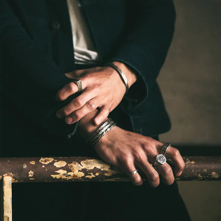 A person wearing several handmade jewellery pieces, including bracelets and rings with unique patterns, crosses their arms over a rusted, weathered metal railing. They are dressed in a dark jacket and light shirt, with the focus on their hands and accessories. One of the standout items is the Nut Ring by Septentrion.