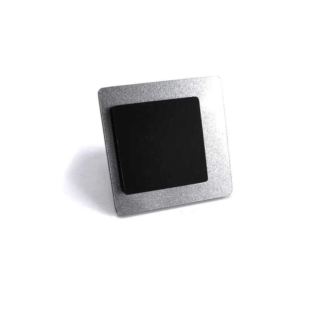 A square, metallic desk plaque with a black surface sits on a white background. The plaque is propped up, slightly tilted backward, displaying a sleek, minimalist design that adds a contemporary touch reminiscent of the Alex + Svet Eriko Ring.