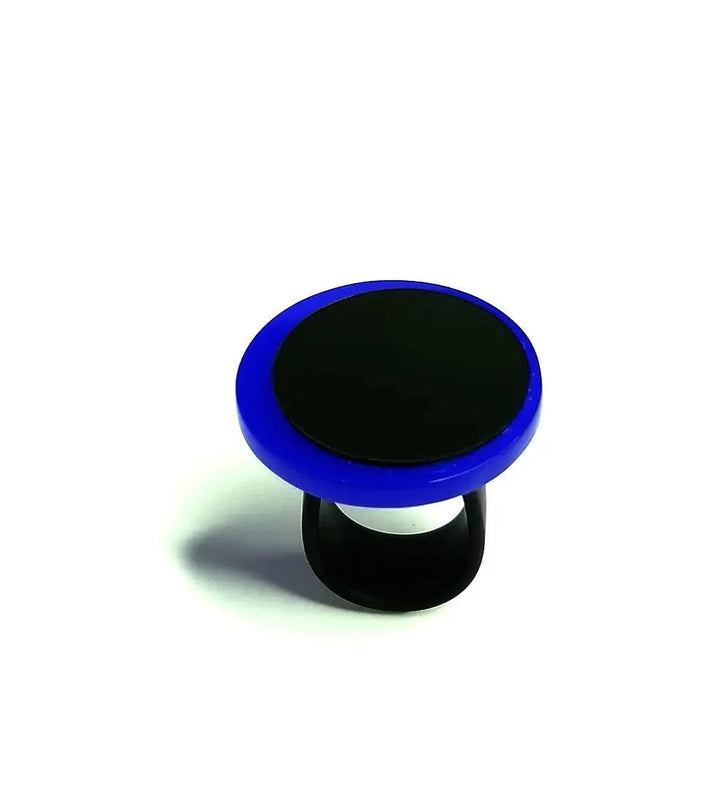 An Amy Ring by Alex + Svet. It features a round, flat top with a blue edge and a black center, reminiscent of an elegant statement ring. The black expandable base attaches to the back of a smartphone, offering both enhanced grip and hands-free use.