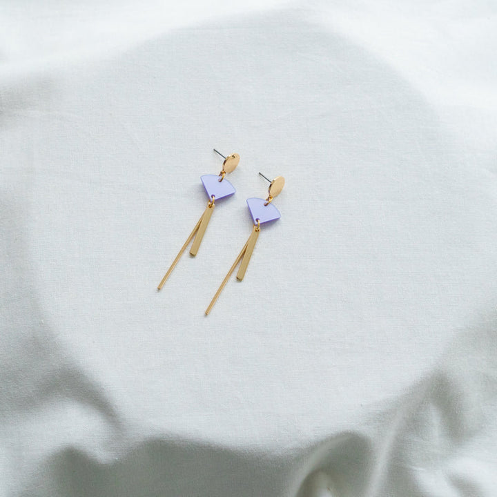 A pair of **Sun 08 Earrings** by **Studio Nok Nok** on a white fabric surface. The **Sun 08 Earrings** feature a triangular stud and a light purple, diamond-shaped piece made from recycled acrylic, connected to two long, slender gold dangling bars. The white fabric has subtle folds and wrinkles.