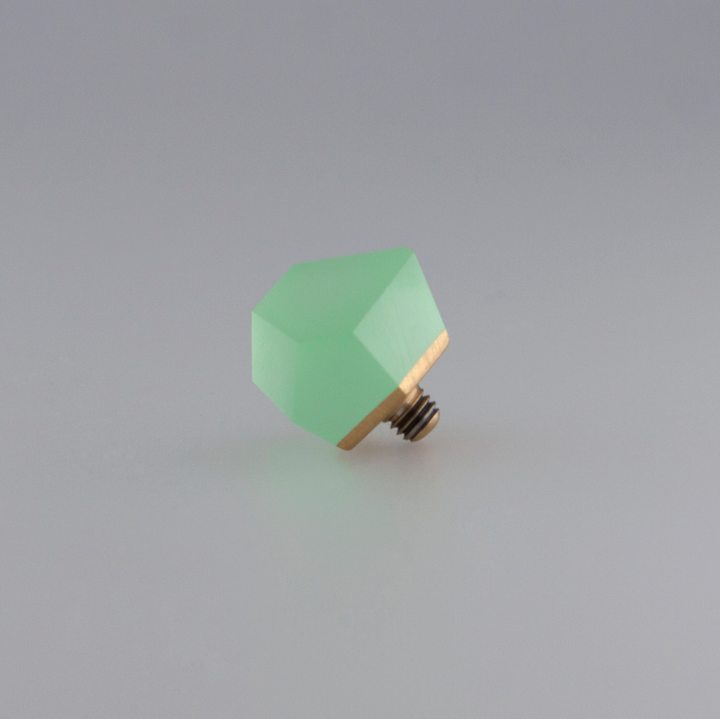 A photo shows a small, faceted, green Fruit Bijoux Ring Top VU Crystals Flash Green knob with a 24ct gold-plated brass base and screw threads at the bottom, placed on a smooth, light gray surface. The gemstone has a slightly translucent quality, giving it a subtle glow.