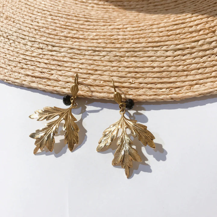 A pair of gold plated brass earrings with intricate leaf-shaped detailing, hanging from small black beads, is displayed against a woven straw surface. The light highlights the metallic sheen of the Ella Maple Leaf Earrings Gold by Lotta et Djossou, emphasizing their elegant and delicate design. Nickel free for added comfort.